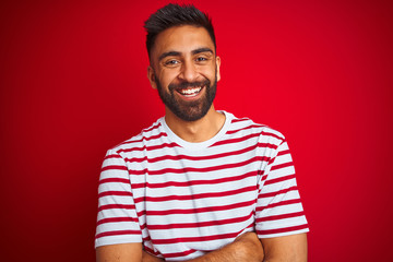 Young indian man wearing striped t-shirt standing over isolated red background happy face smiling with crossed arms looking at the camera. Positive person.