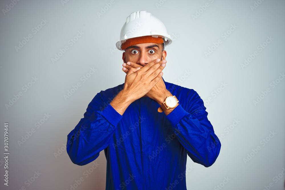 Handsome indian worker man wearing uniform and helmet over isolated white background shocked covering mouth with hands for mistake. Secret concept.