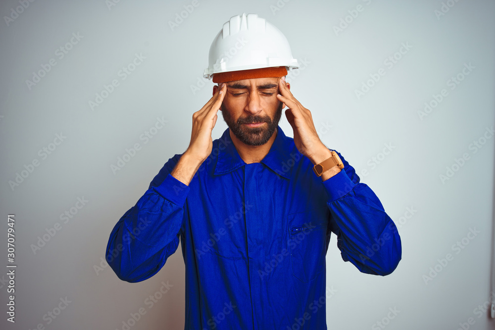 Handsome indian worker man wearing uniform and helmet over isolated white background with hand on head for pain in head because stress. Suffering migraine.