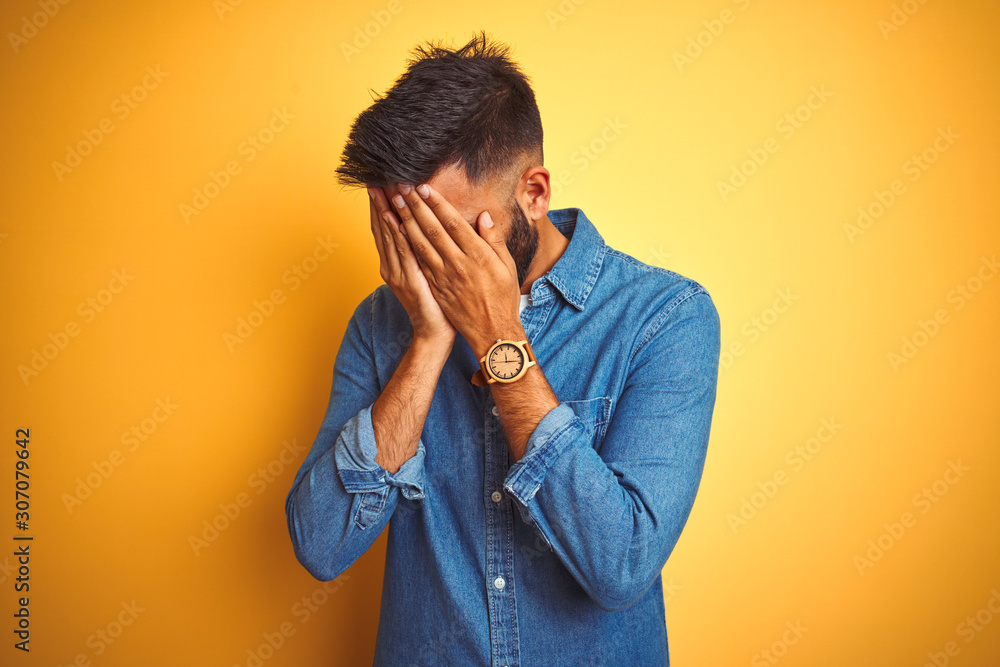 Young indian man wearing denim shirt standing over isolated yellow background with sad expression covering face with hands while crying. Depression concept. Young indian man wearing denim shirt standing over isolated yellow background with sad expression covering face with hands while crying. Depression concept.