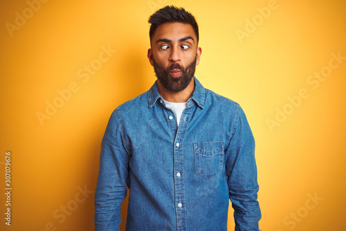 Young indian man wearing denim shirt standing over isolated yellow background making fish face with lips, crazy and comical gesture. Funny expression.