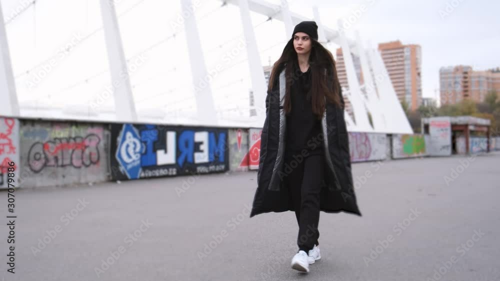 Fashion portrait of trendy pretty brunette girl walking outdoors. Looking at the camera. Female model wearing stylish long black jacket 4k Fashion portrait of trendy pretty brunette girl walking outdoors. Looking at the camera. Female model wearing stylish long black jacket 4k