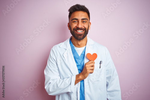 Young indian doctor man holding paper heart standing over isolated pink background with a happy face standing and smiling with a confident smile showing teeth © Krakenimages.com