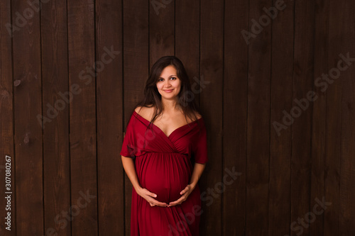 Christmas pregnant woman in red dress holds hands on belly on a dark brown background. Pregnancy, maternity, expectation concept. Beautiful tender mood photo of pregnancy