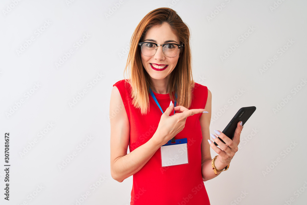 Naklejka premium Redhead businesswoman wearing id card using smartphone over isolated white background very happy pointing with hand and finger