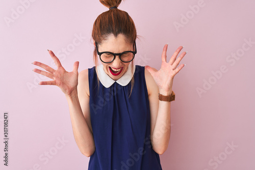 Redhead woman with pigtail wearing elegant dress and glasses over isolated pink background celebrating mad and crazy for success with arms raised and closed eyes screaming excited. Winner concept