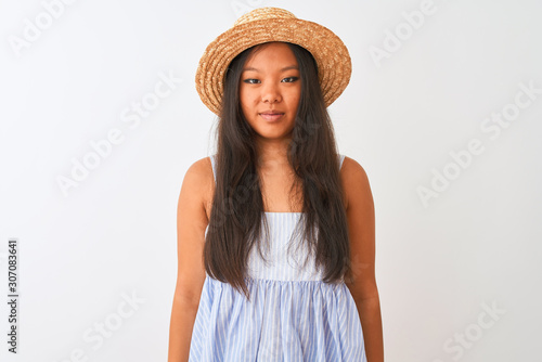 Young chinese woman wearing striped dress and hat standing over isolated white background with serious expression on face. Simple and natural looking at the camera.