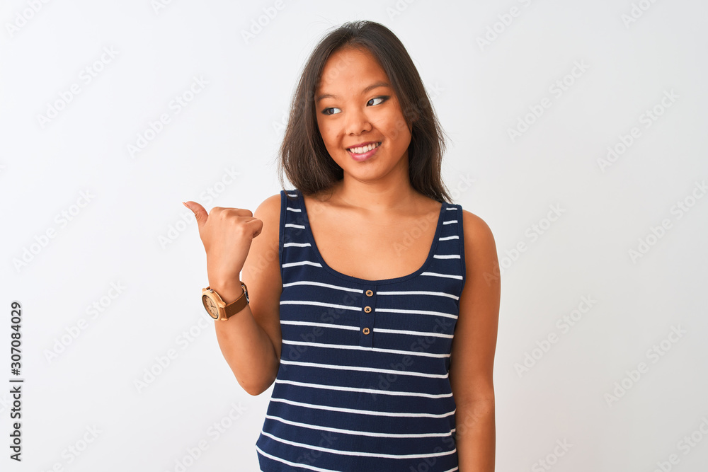 Young chinese woman wearing striped t-shirt standing over isolated white background smiling with happy face looking and pointing to the side with thumb up.