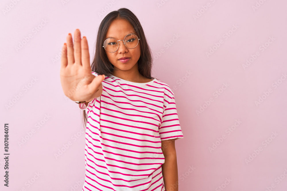 Young chinese woman wearing striped t-shirt and glasses over isolated pink background doing stop sing with palm of the hand. Warning expression with negative and serious gesture on the face. Young chinese woman wearing striped t-shirt and glasses over isolated pink background doing stop sing with palm of the hand. Warning expression with negative and serious gesture on the face.
