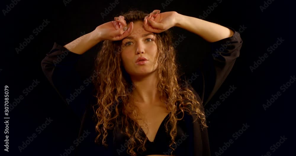 A close-up portrait of a smiling curly-haired young girl who is in the Studio on a black background, wearing a black unbuttoned jacket and a black bra, she poses and touches her hair, dances. A close-up portrait of a smiling curly-haired young girl who is in the Studio on a black background, wearing a black unbuttoned jacket and a black bra, she poses and touches her hair, dances.