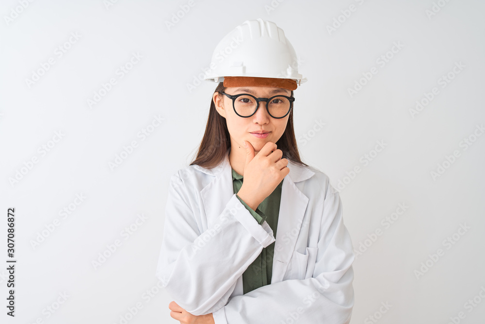 Young chinese engineer woman wearing coat helmet glasses over isolated white background looking confident at the camera with smile with crossed arms and hand raised on chin. Thinking positive. Young chinese engineer woman wearing coat helmet glasses over isolated white background looking confident at the camera with smile with crossed arms and hand raised on chin. Thinking positive.