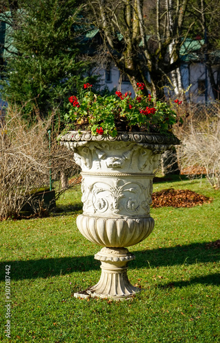 Decorative stone flowerpot with floral and artistic incrustations and on top bouquet of small red blossomed flowers , inside a garden