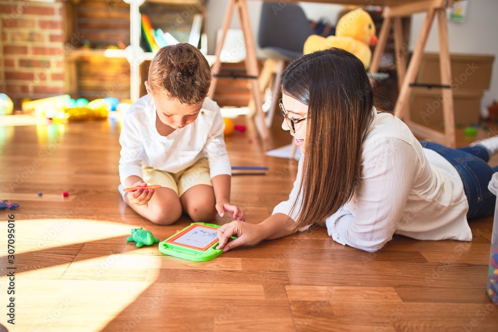 Fototapeta premium Beautiful teacher and toddler drawing using digital board around lots of toys at kindergarten