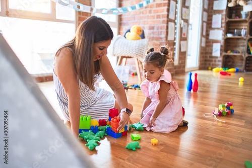 Young beautiful teacher and toddler sitting on the floor playing with building blocks toy at kindergarten
