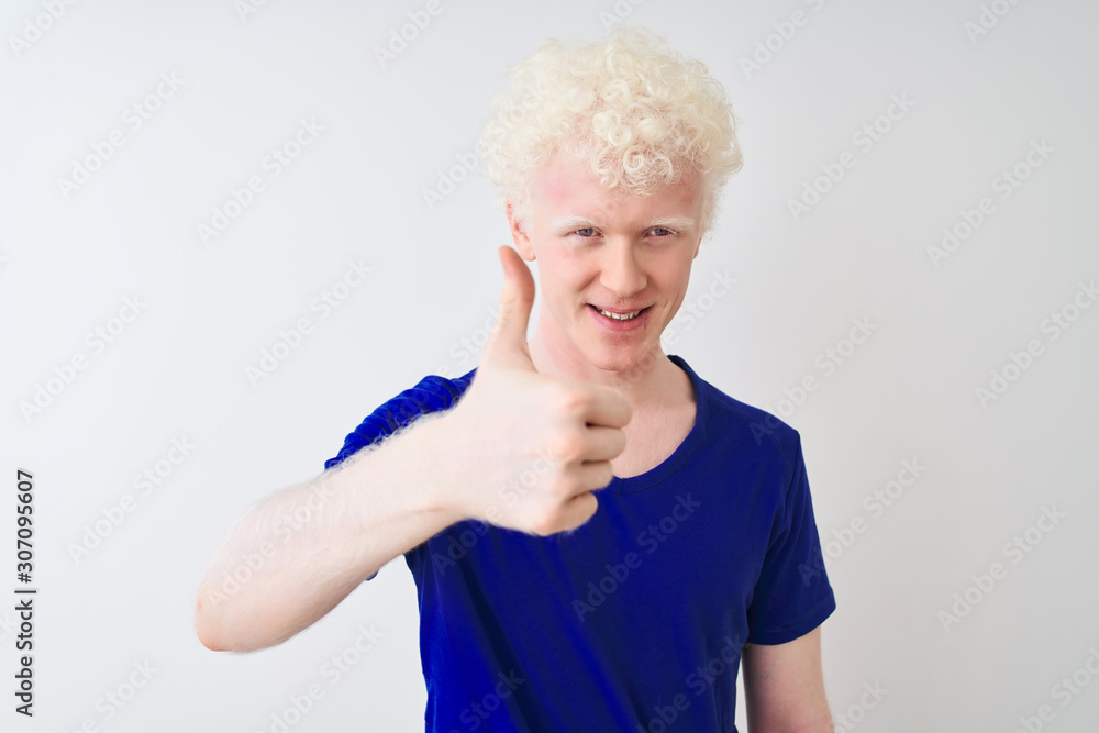 Young albino blond man wearing blue casual t-shirt standing over isolated white background doing happy thumbs up gesture with hand. Approving expression looking at the camera with showing success. Young albino blond man wearing blue casual t-shirt standing over isolated white background doing happy thumbs up gesture with hand. Approving expression looking at the camera with showing success.