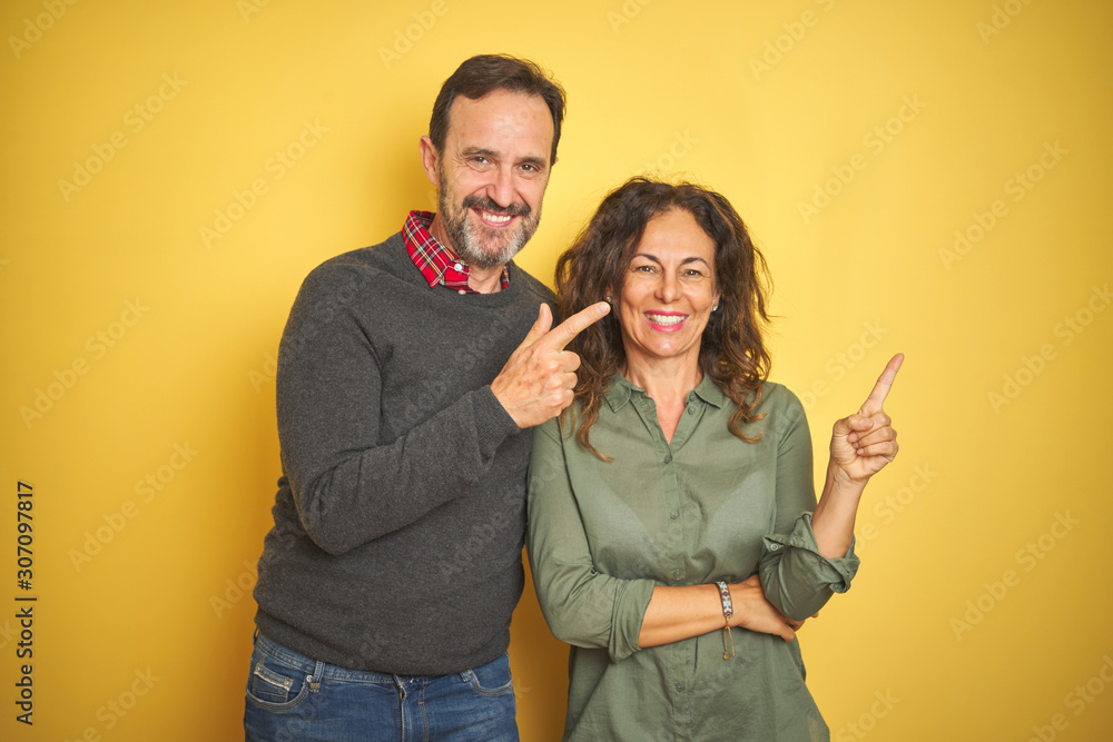Beautiful middle age couple over isolated yellow background with a big smile on face, pointing with hand and finger to the side looking at the camera. Beautiful middle age couple over isolated yellow background with a big smile on face, pointing with hand and finger to the side looking at the camera.