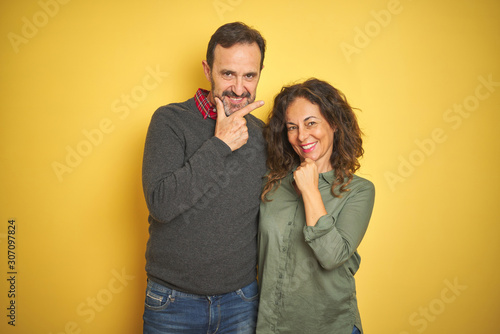 Beautiful middle age couple over isolated yellow background looking confident at the camera with smile with crossed arms and hand raised on chin. Thinking positive.