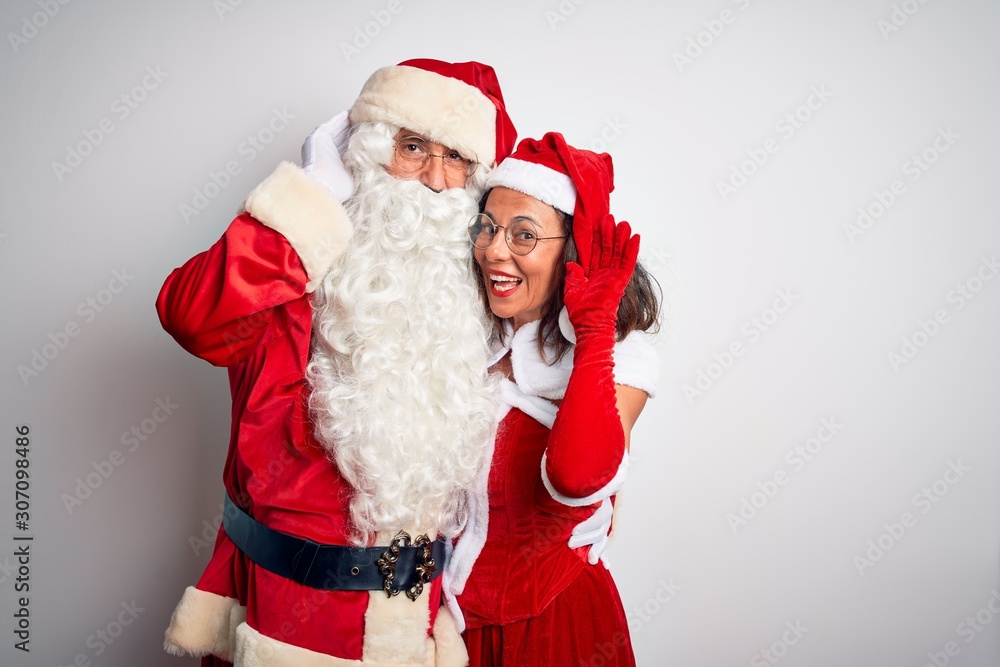 Middle age couple wearing Santa costume hugging over isolated white background smiling with hand over ear listening an hearing to rumor or gossip. Deafness concept. Middle age couple wearing Santa costume hugging over isolated white background smiling with hand over ear listening an hearing to rumor or gossip. Deafness concept.