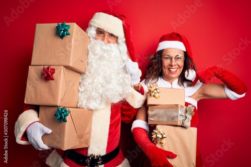 Middle age couple wearing Santa costume holding tower of gifts over isolated red background looking confident with smile on face, pointing oneself with fingers proud and happy.