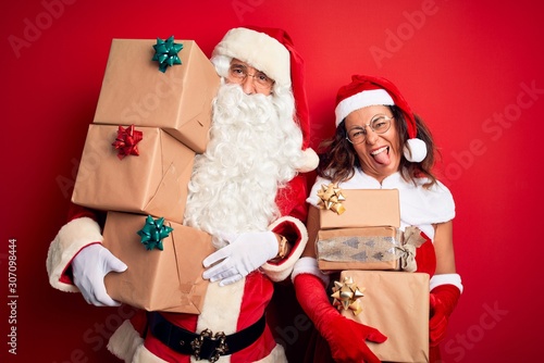 Middle age couple wearing Santa costume holding tower of gifts over isolated red background sticking tongue out happy with funny expression. Emotion concept.