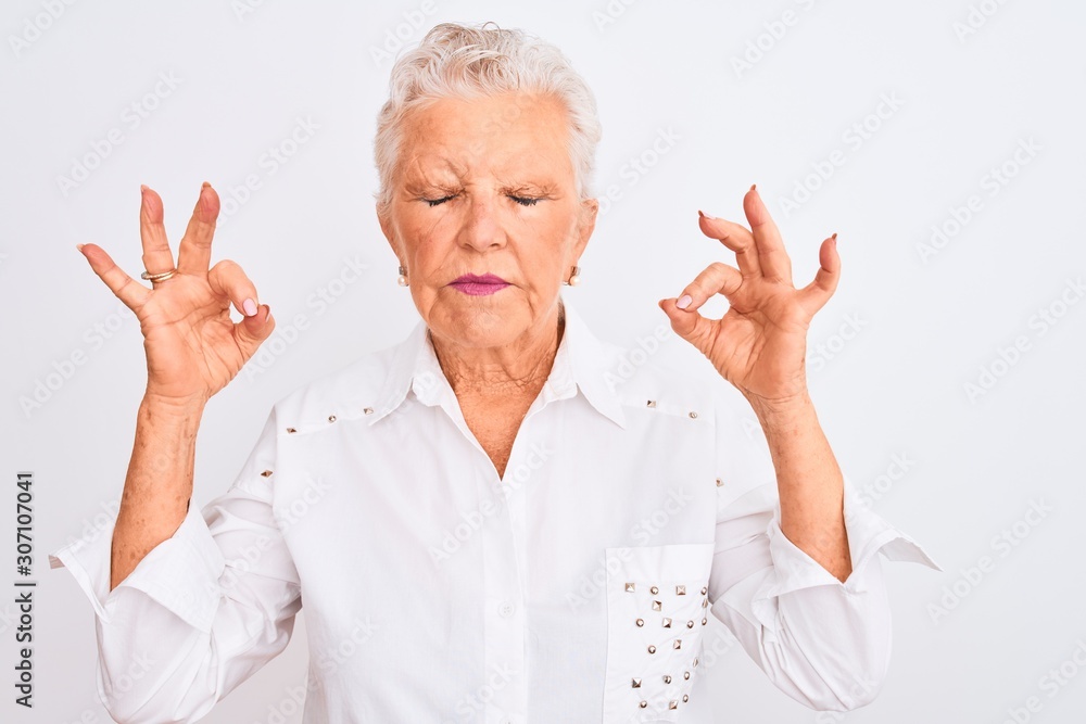 Senior grey-haired woman wearing elegant shirt standing over isolated white background relax and smiling with eyes closed doing meditation gesture with fingers. Yoga concept. Senior grey-haired woman wearing elegant shirt standing over isolated white background relax and smiling with eyes closed doing meditation gesture with fingers. Yoga concept.