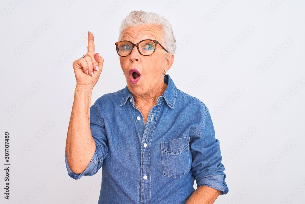 Senior grey-haired woman wearing denim shirt and glasses over isolated white background pointing finger up with successful idea. Exited and happy. Number one. Senior grey-haired woman wearing denim shirt and glasses over isolated white background pointing finger up with successful idea. Exited and happy. Number one.
