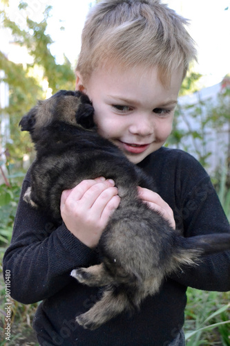 A little boy in a black turtleneck hugs a black puppy and smiles
