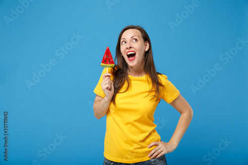 Pensive pretty young brunette woman girl in yellow t-shirt posing isolated on bright blue background studio portrait. People lifestyle concept. Mock up copy space. Hold watermelon lollipop looking up. © ViDi Studio
