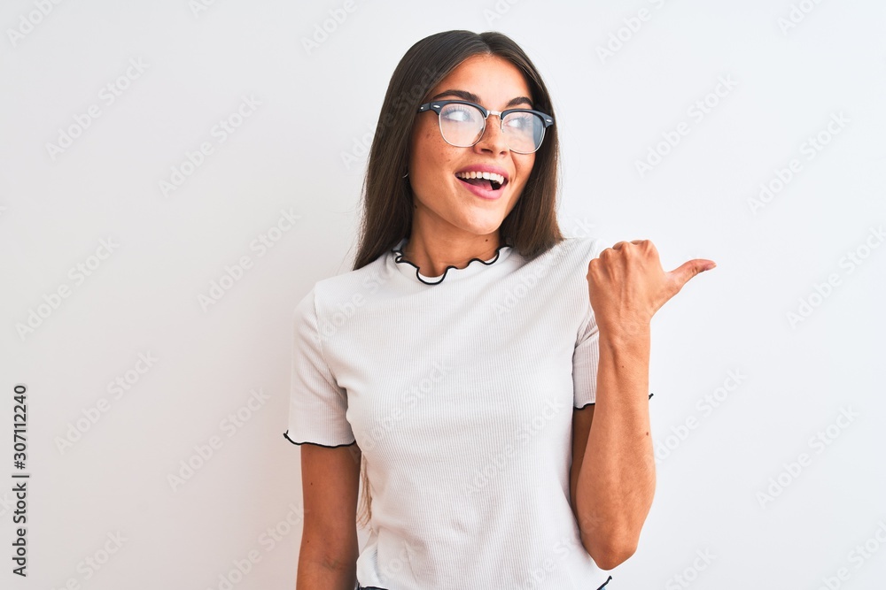 Young beautiful woman wearing casual t-shirt and glasses over isolated white background smiling with happy face looking and pointing to the side with thumb up. Young beautiful woman wearing casual t-shirt and glasses over isolated white background smiling with happy face looking and pointing to the side with thumb up.