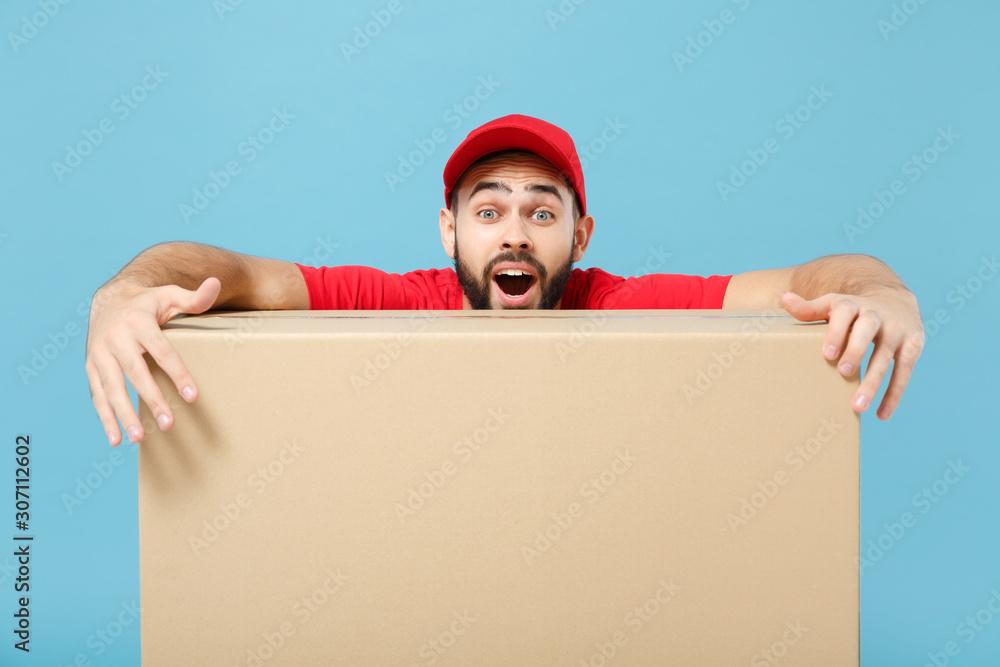 Delivery man in red uniform isolated on blue background, studio portrait. Male employee in cap t-shirt print working as courier dealer hold empty cardboard box. Service concept. Mock up copy space. Delivery man in red uniform isolated on blue background, studio portrait. Male employee in cap t-shirt print working as courier dealer hold empty cardboard box. Service concept. Mock up copy space.