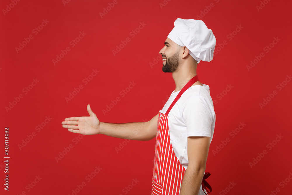 Side view of young male chef cook or baker man in striped apron toque chefs hat posing isolated on red background. Cooking food concept. Mock up copy space. Stand with outstretched hand for greeting. Side view of young male chef cook or baker man in striped apron toque chefs hat posing isolated on red background. Cooking food concept. Mock up copy space. Stand with outstretched hand for greeting.
