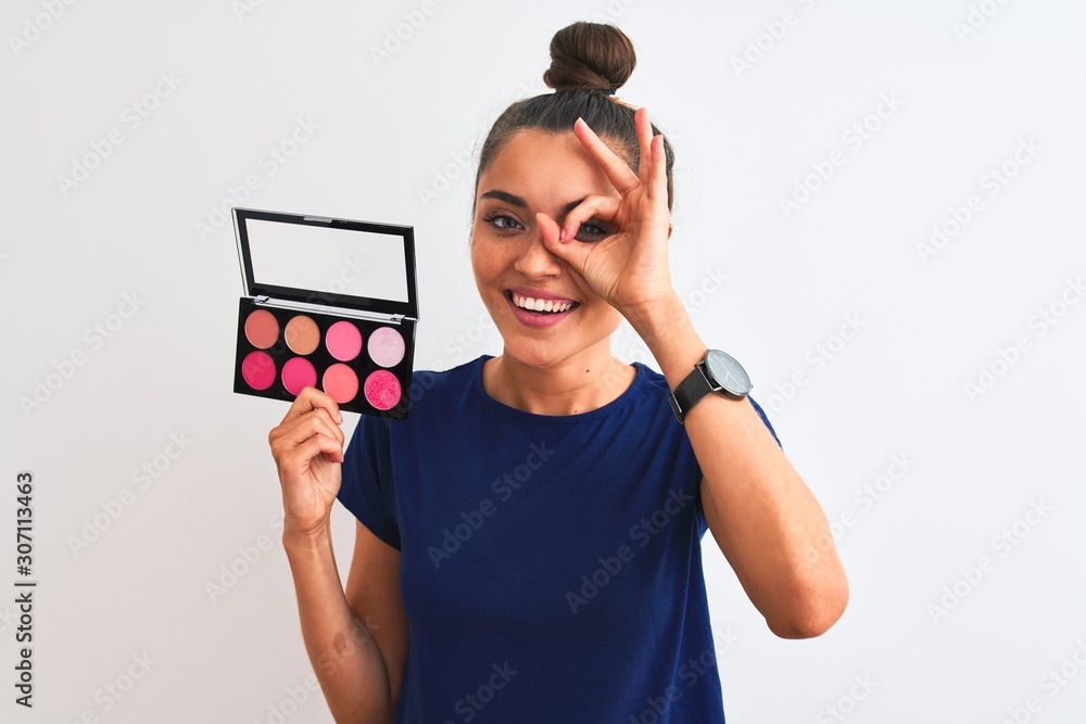 Young beautiful woman holding makeup standing over isolated white background with happy face smiling doing ok sign with hand on eye looking through fingers Young beautiful woman holding makeup standing over isolated white background with happy face smiling doing ok sign with hand on eye looking through fingers
