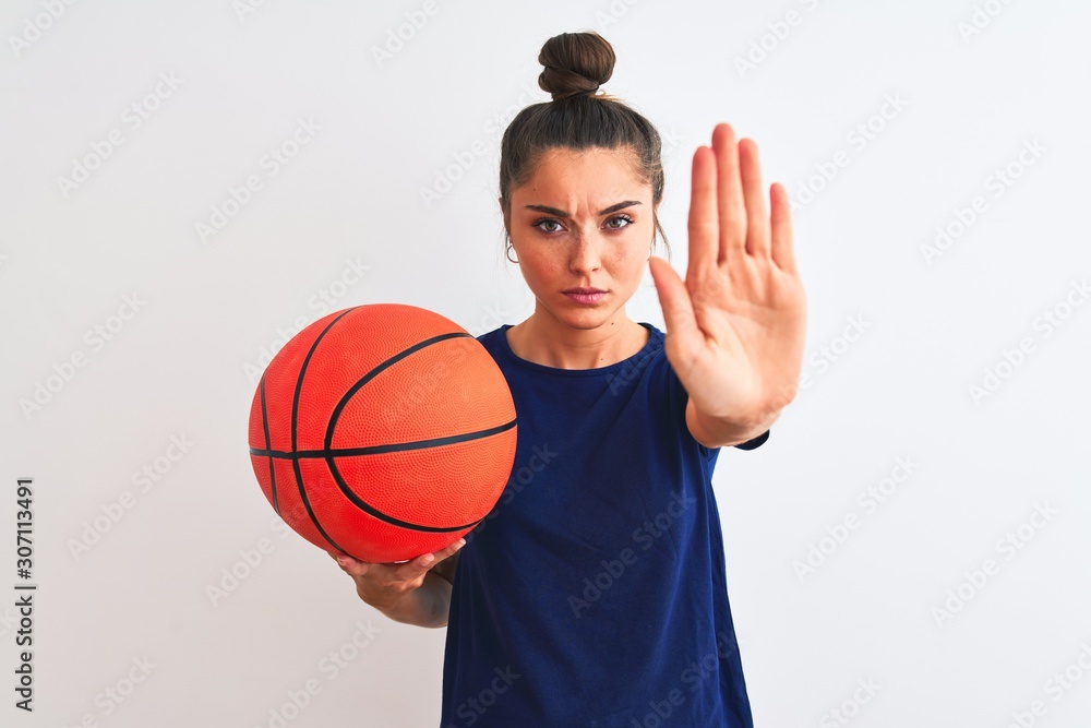 Young beautiful sportswoman holding basketball ball over isolated white background with open hand doing stop sign with serious and confident expression, defense gesture