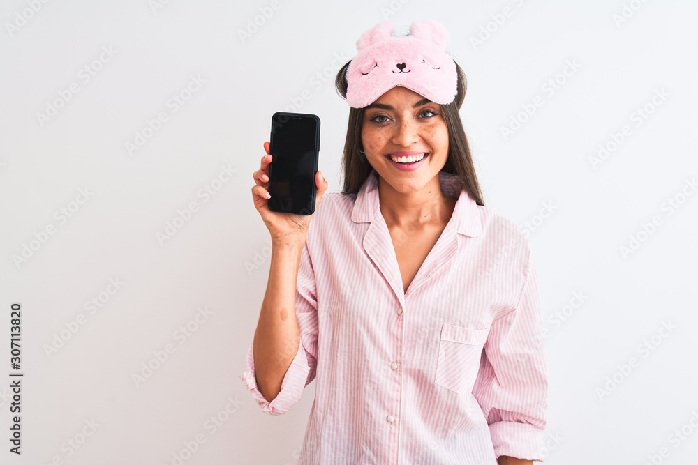 Young woman wearing sleep mask pajama holding smartphone over isolated white background with a happy face standing and smiling with a confident smile showing teeth Young woman wearing sleep mask pajama holding smartphone over isolated white background with a happy face standing and smiling with a confident smile showing teeth