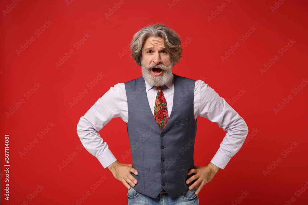Irritated elderly gray-haired mustache bearded man in classic shirt vest colorful tie isolated on red wall background. People lifestyle concept. Mock up copy space. Standing with arms akimbo swearing. Irritated elderly gray-haired mustache bearded man in classic shirt vest colorful tie isolated on red wall background. People lifestyle concept. Mock up copy space. Standing with arms akimbo swearing.