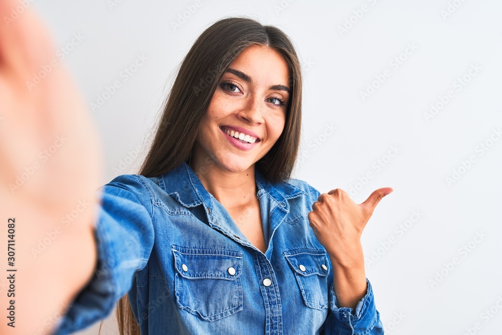 Beautiful woman wearing denim shirt make selfie by camera over isolated white background Pointing to the back behind with hand and thumbs up, smiling confident Beautiful woman wearing denim shirt make selfie by camera over isolated white background Pointing to the back behind with hand and thumbs up, smiling confident