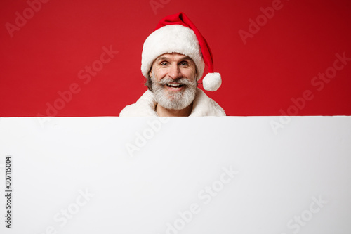 Smiling elderly gray-haired mustache bearded Santa man in Christmas hat posing isolated on red background. New Year 2020 celebration holiday concept. Mock up copy space. Holding blank white billboard.