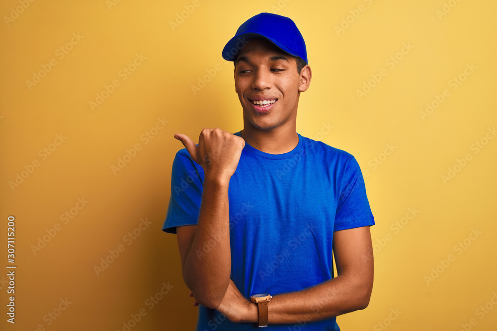 Young handsome arab delivery man standing over isolated yellow background smiling with happy face looking and pointing to the side with thumb up.