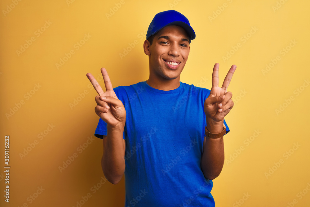 Young handsome arab delivery man standing over isolated yellow background smiling looking to the camera showing fingers doing victory sign. Number two.