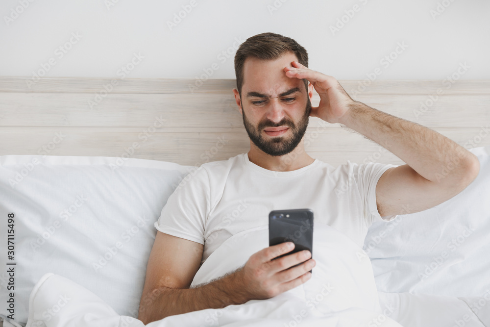 Calm young handsome bearded man lying in bed with white sheet pillow blanket in bedroom at home. Male spending time in room with mobile phone. Rest relax good mood lifestyle concept Mock up copy space Calm young handsome bearded man lying in bed with white sheet pillow blanket in bedroom at home. Male spending time in room with mobile phone. Rest relax good mood lifestyle concept Mock up copy space