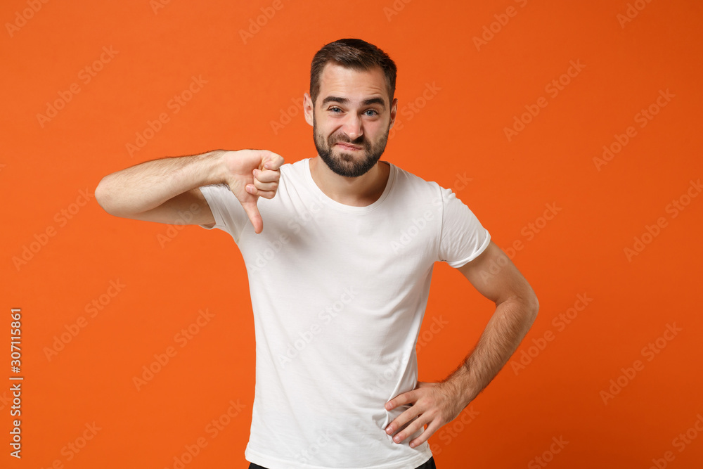 Naklejka premium Displeased confused young man in casual white t-shirt posing isolated on orange wall background studio portrait. People sincere emotions lifestyle concept. Mock up copy space. Showing thumb down.