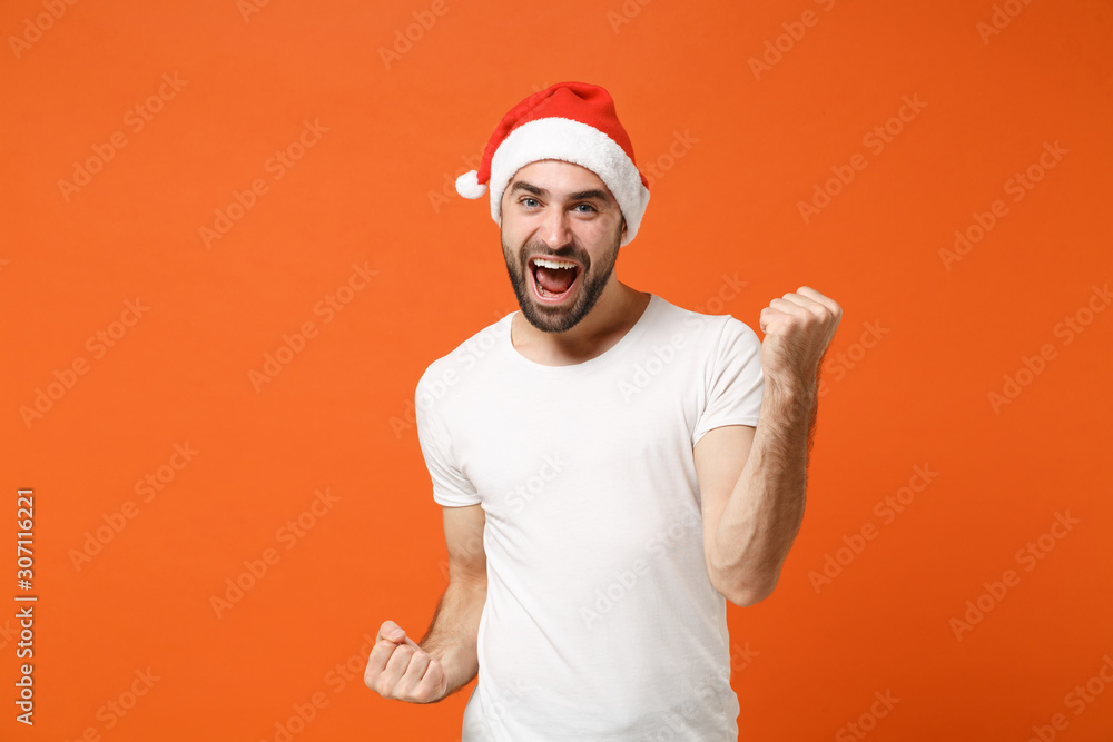 Happy young Santa man in white t-shirt, Christmas hat posing isolated on orange background in studio. Happy New Year 2020 celebration holiday concept. Mock up copy space. Clenching fists like winner.