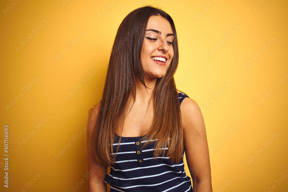 Young beautiful woman wearing striped t-shirt standing over isolated yellow background looking away to side with smile on face, natural expression. Laughing confident. Young beautiful woman wearing striped t-shirt standing over isolated yellow background looking away to side with smile on face, natural expression. Laughing confident.