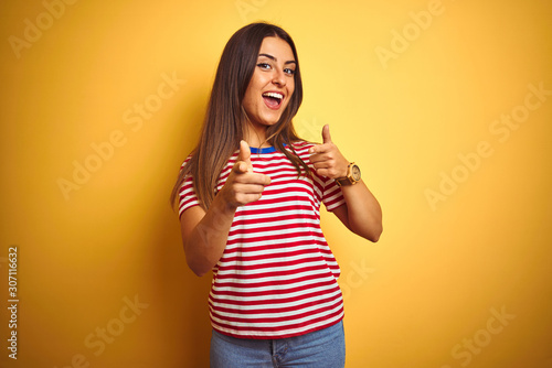Young beautiful woman wearing striped t-shirt standing over isolated yellow background pointing fingers to camera with happy and funny face. Good energy and vibes.