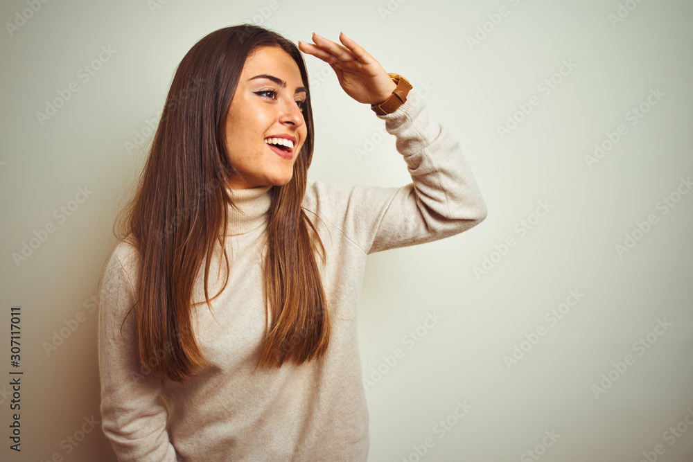 Young beautiful woman wearing winter turtleneck sweater over isolated white background very happy and smiling looking far away with hand over head. Searching concept. Young beautiful woman wearing winter turtleneck sweater over isolated white background very happy and smiling looking far away with hand over head. Searching concept.