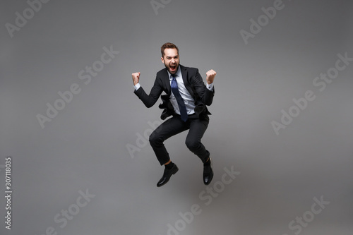 Joyful young business man in classic black suit shirt tie posing isolated on grey background. Achievement career wealth business concept. Mock up copy space. Jumping, doing winner gesture, screaming.