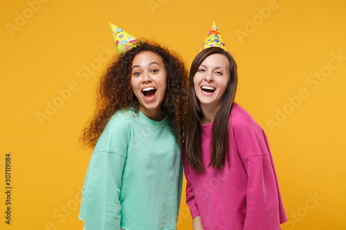 Two excited laughing women friends european and african american girls in pink green clothes, birthday hats posing isolated on yellow orange background. People lifestyle concept. Mock up copy space.