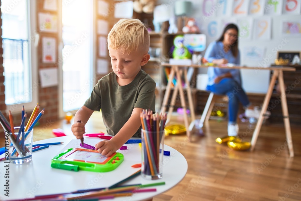 Young caucasian child playing at playschool with teacher. Mother and son at playroom drawing a draw with color pencils, young woman at the background sitting on desk. Young caucasian child playing at playschool with teacher. Mother and son at playroom drawing a draw with color pencils, young woman at the background sitting on desk.