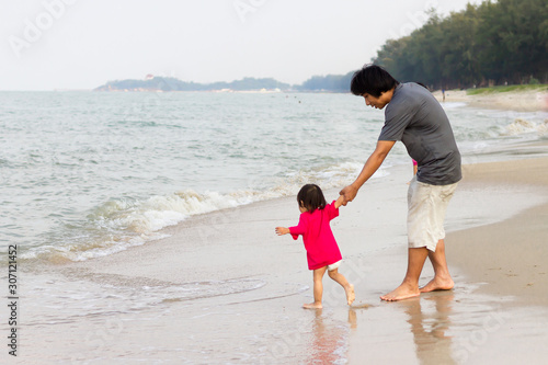 Portrait image of Asian Father with his daughter are​ hand​ in​ hand​ and​ walking​ on the beach at the sea. Summer season. Father's day and family concept.