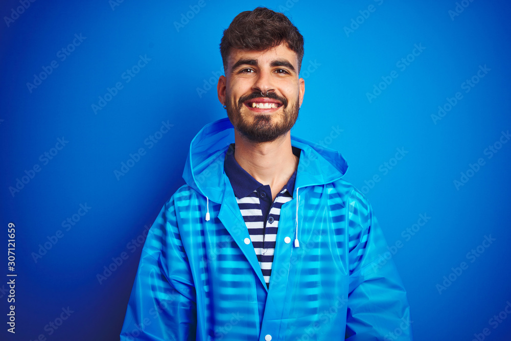 Naklejka premium Young handsome man wearing rain coat standing over isolated blue background with a happy and cool smile on face. Lucky person.